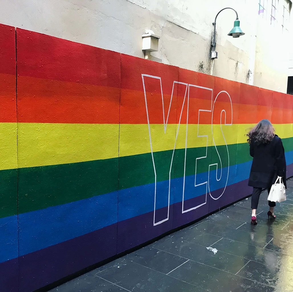 Person walking past a large rainbow wall mural with the word YES painted on it